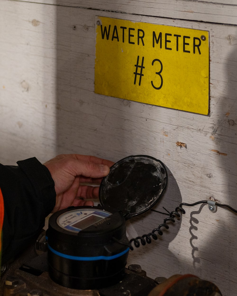 A worker in high-visibility clothing lifting the lid on a water meter, beside a Quantify enclosure